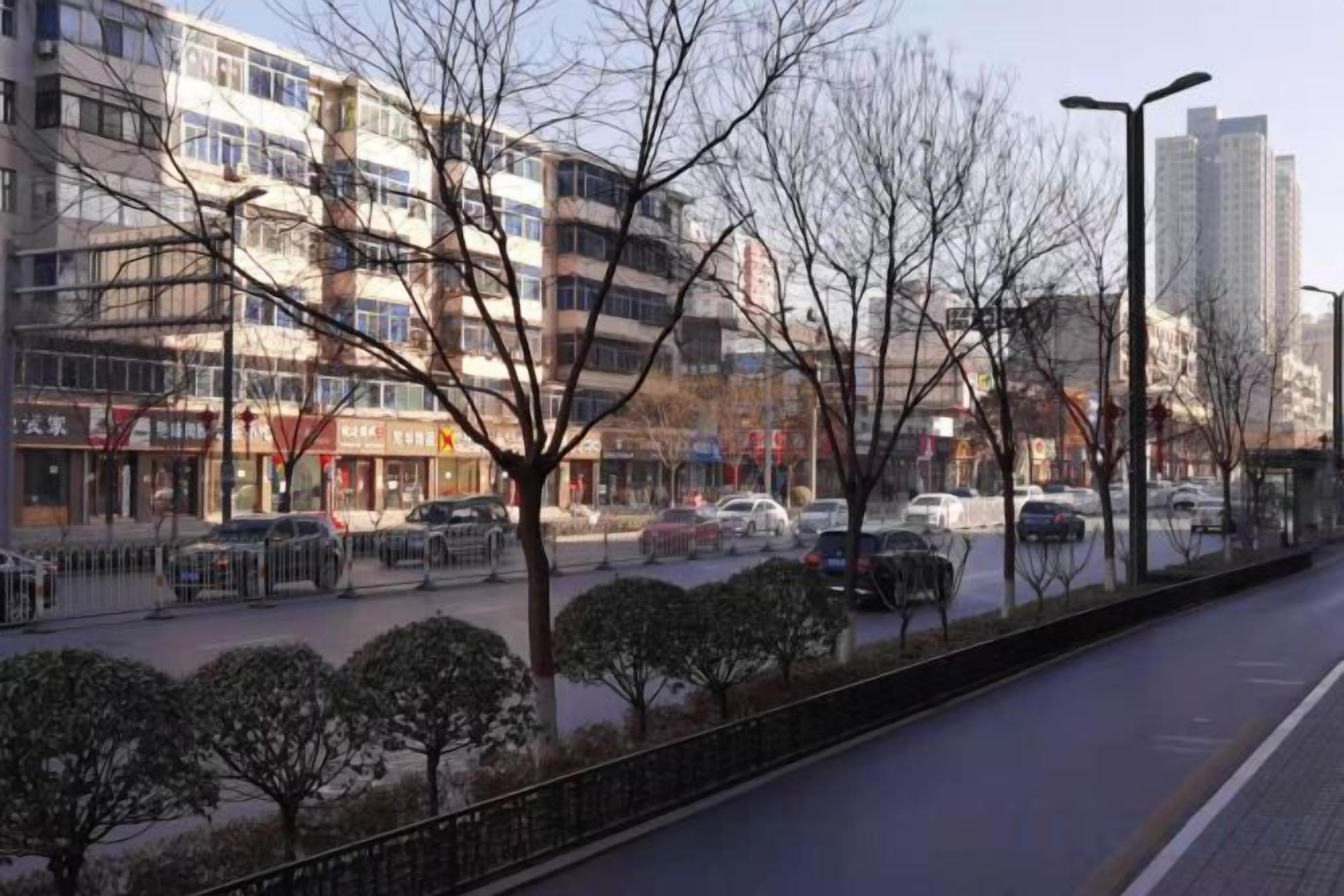 A multi-story building with a row of shops on the ground floor, a parking lot with cars, and a sidewalk with trees.