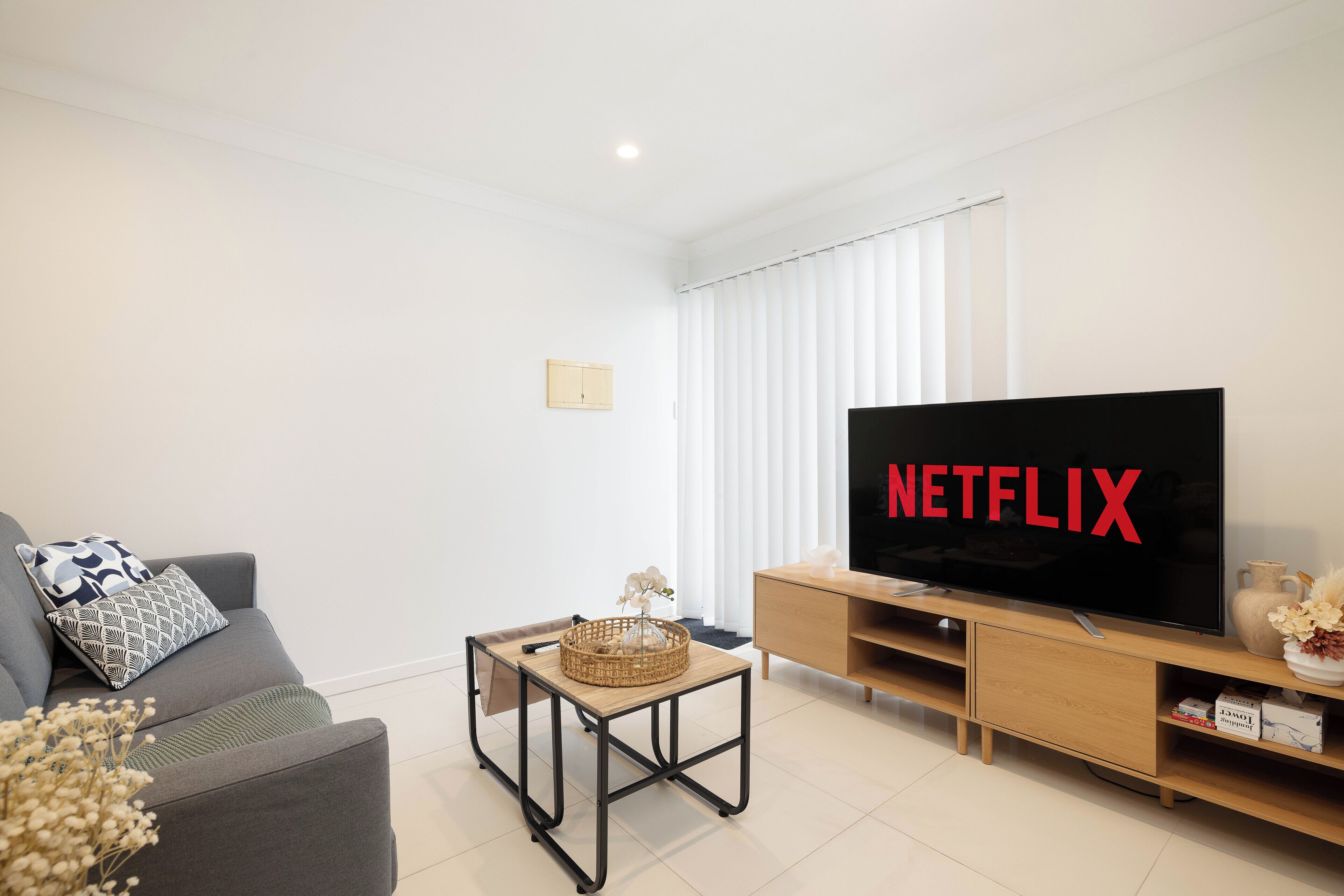 A modern living room with a grey sofa, a coffee table with a woven basket, a wooden TV stand with a flat-screen TV displaying "NETFLIX", and a light wooden cabinet with a vase.