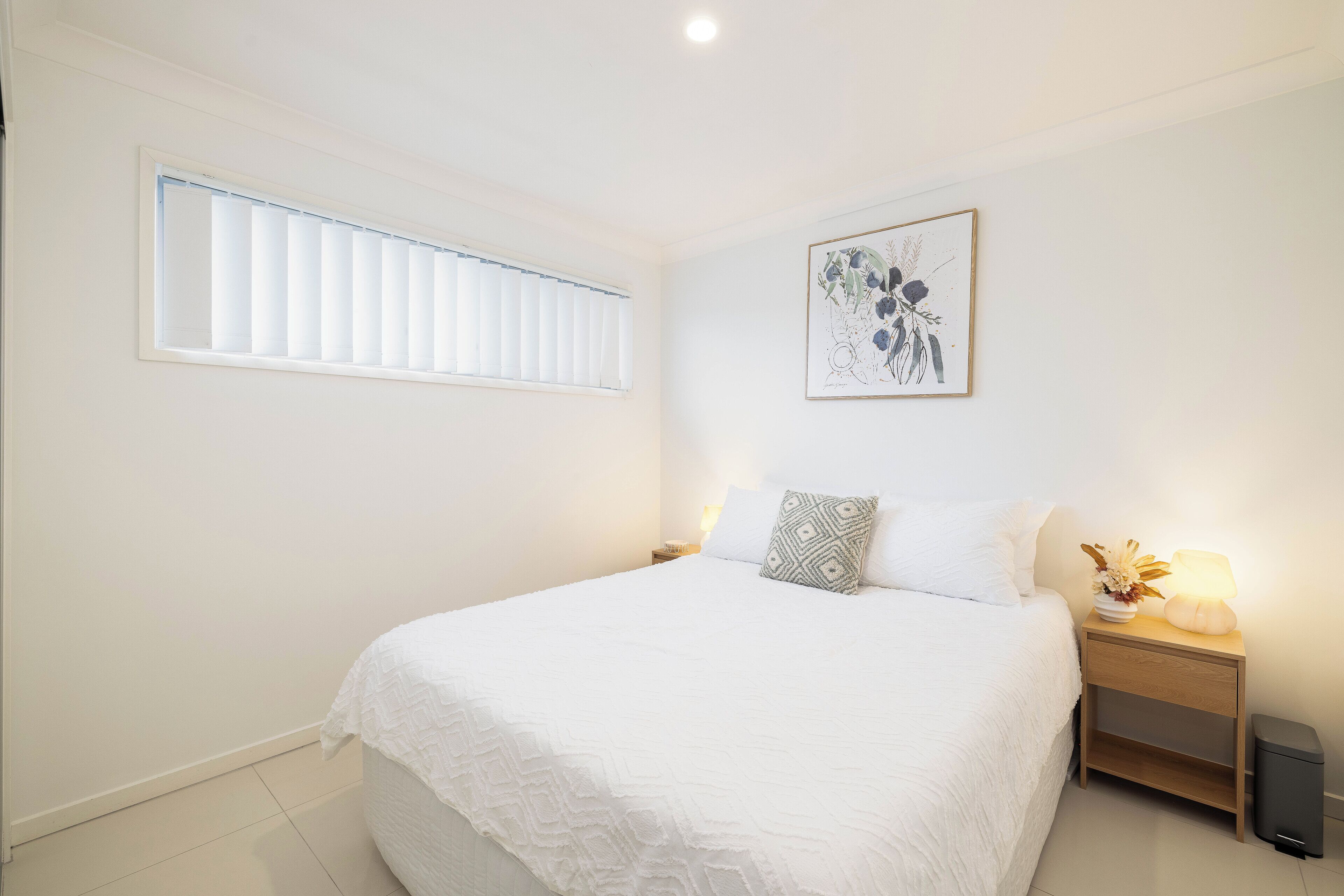 A bedroom with a bed, a nightstand with a lamp, a framed artwork on the wall, and a window with blinds.