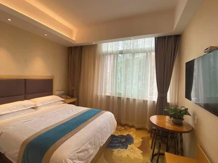 Ein Hotelzimmer mit einem Bett, einem Fernseher, einem kleinen Tisch mit Pflanze und einem Fenster mit Vorhängen.