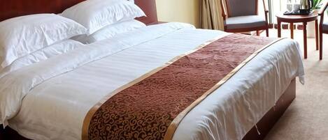 A neatly made bed with white linens and a patterned throw, a wooden headboard, and a small table with a plant beside the bed.