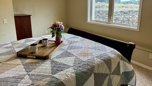 A bedroom with a large bed, a window with a view of a rocky landscape, a wooden nightstand with a vase of flowers, and a tray with two mugs.