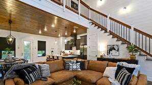 A spacious living area with a brown leather sectional sofa, a wooden staircase, and a kitchen in the background.