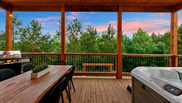 A wooden deck with a hot tub, barbecue grill, and seating area overlooking a forested landscape.