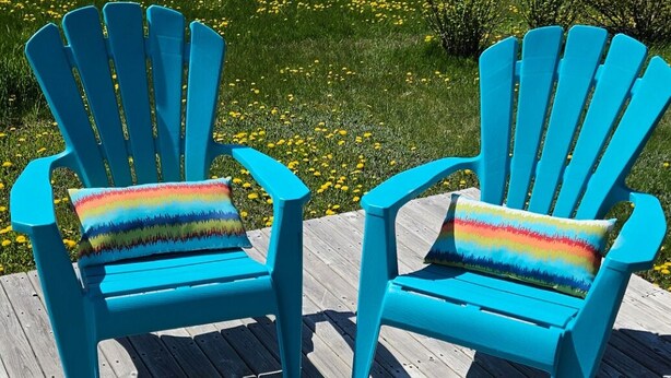 To turkise Adirondack-stole med farverige hynder på en træterrasse med en smuk udsigt over marker og træer.