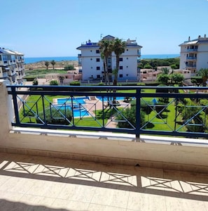 A balcony with a view of a swimming pool, a building with a white facade, and a clear blue sky.