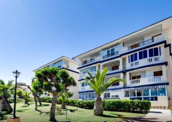 Un hotel de varios pisos con balcones, un jardín bien cuidado y un cielo azul despejado.