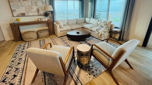 A modern living room with a sectional sofa, two armchairs, a coffee table, and a rug. There is a large window with curtains, a ceiling fan, and a wall-mounted TV.