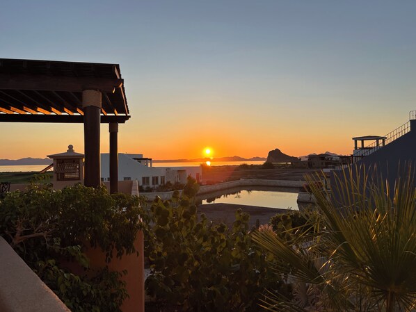 A sunset view from a terrace with a pool, overlooking a body of water and distant mountains.