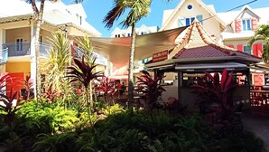 Un restaurante con terraza al aire libre, rodeado de exuberante vegetación y palmeras, con vista a un edificio de techo rojo y balcones.