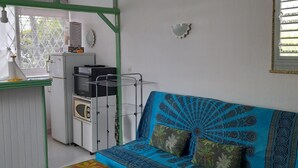 Una habitación con un sofá azul, una mesa blanca, una kitchenette y una ventana con persianas.
