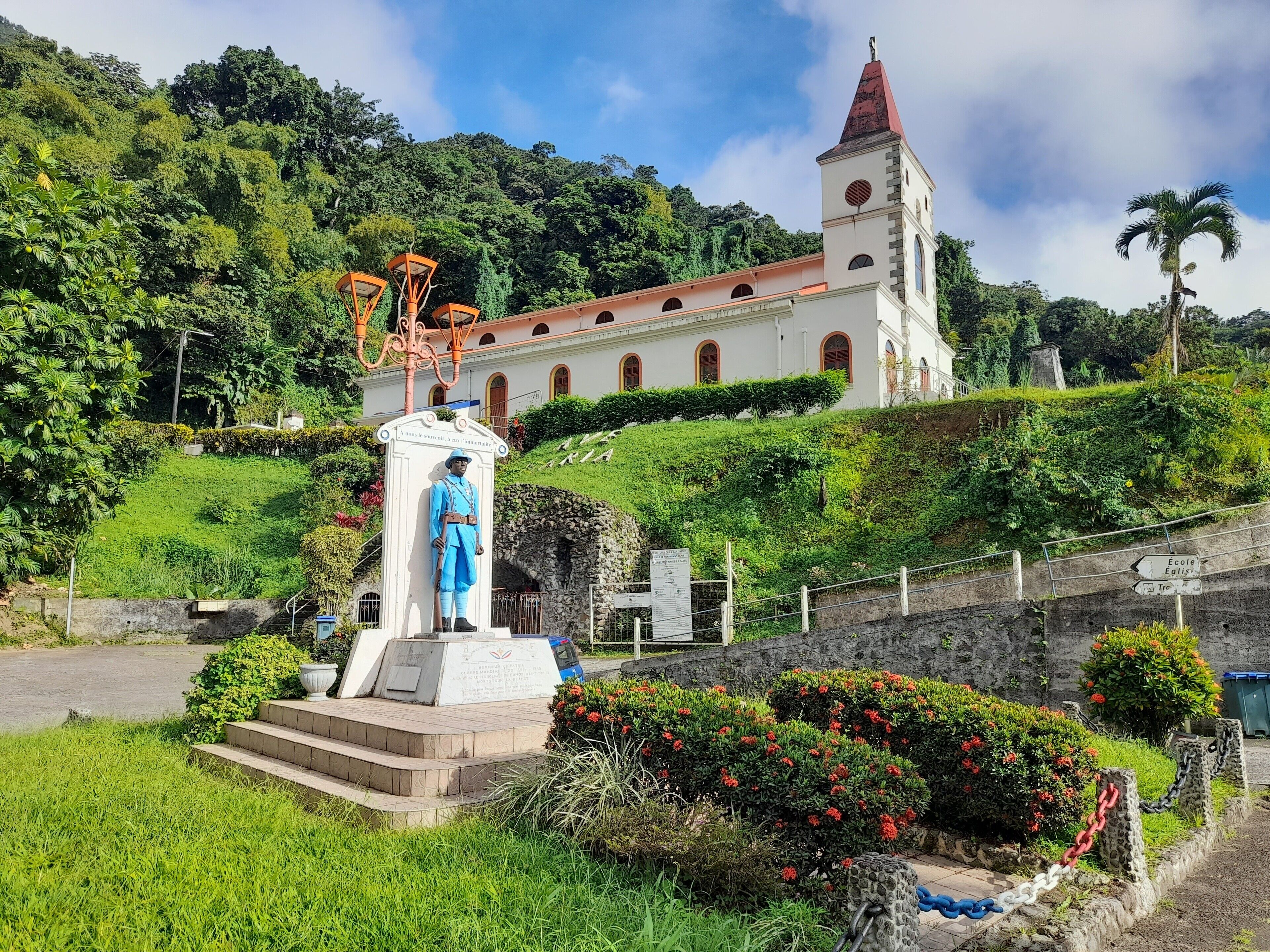 A statue in front of a church with a bell tower, surrounded by greenery and a stone wall with a chain barrier.