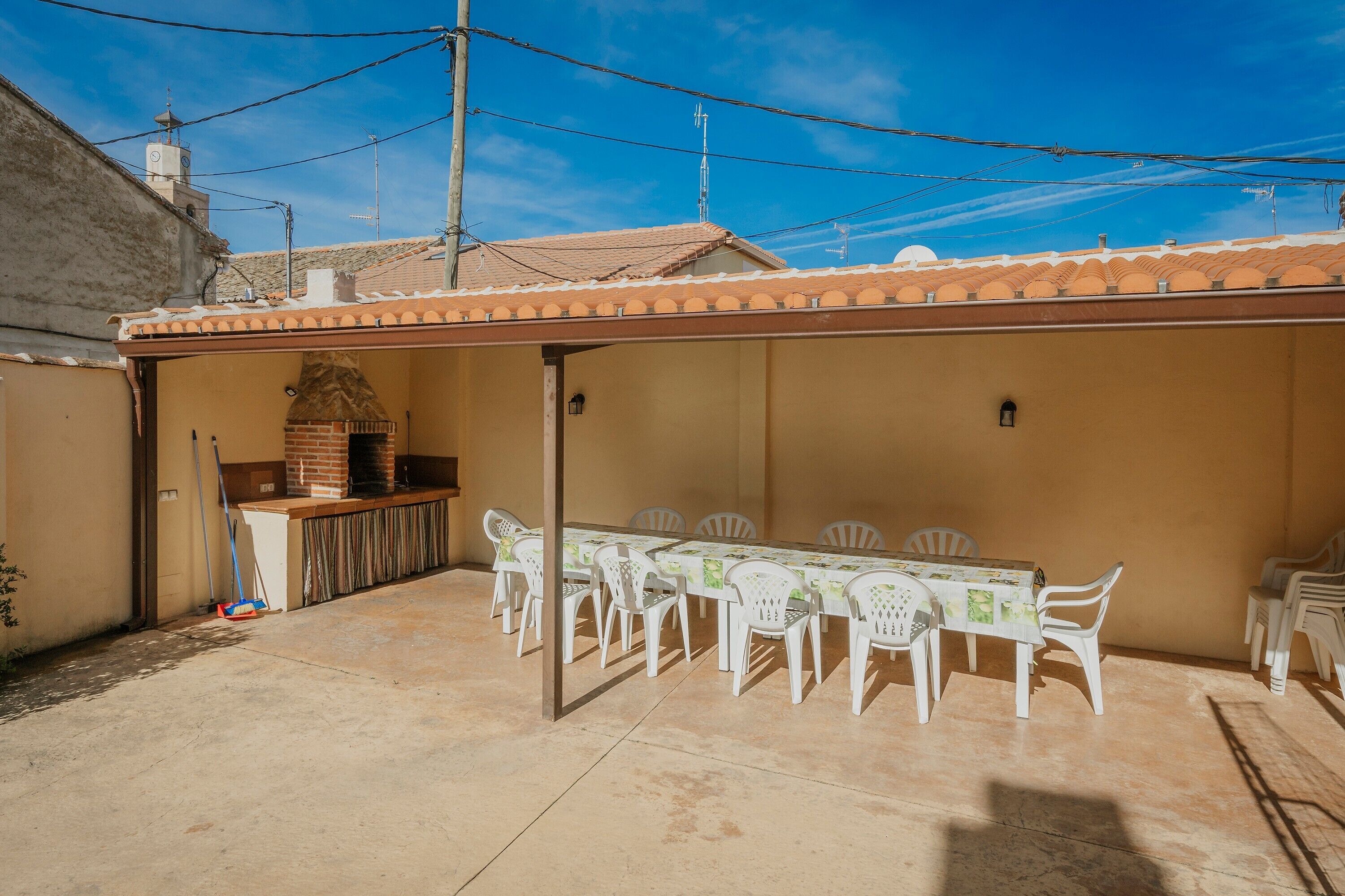 An outdoor patio with a brick oven, white plastic chairs, and a long table.