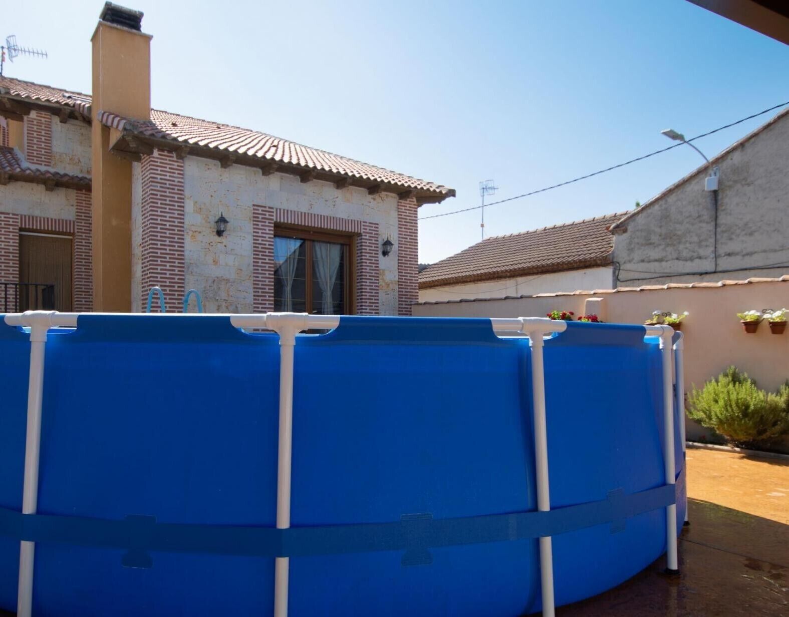 A blue swimming pool with white pipes, a brick building with a tiled roof, and a concrete wall with hanging plants.
