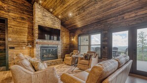 A cozy living room with leather sofas, a stone fireplace, and large windows offering a view of the outdoors. - Get away and relax at Elk Ridge, a modern lodge in the mountains of Ruidoso. (Ruidoso)