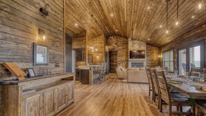 A rustic interior with wooden walls and ceiling, a stone fireplace, a dining area with a large table and chairs, and a seating area with sofas. - Get away and relax at Elk Ridge, a modern lodge in the mountains of Ruidoso. (Ruidoso)