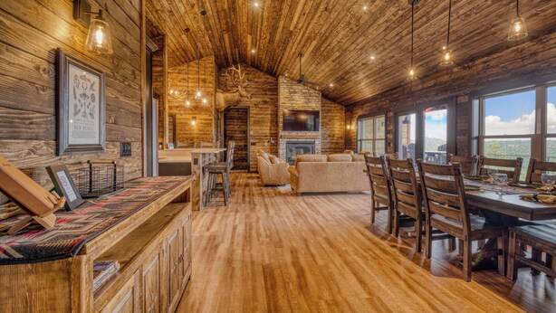 A rustic interior with wooden walls, a stone fireplace, and a dining area with a long table and chairs. - Get away and relax at Elk Ridge, a modern lodge in the mountains of Ruidoso. (Ruidoso)