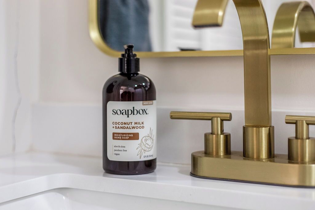 A bathroom with a white sink, a gold faucet, and a soap dispenser labeled 'soapbox Coconut Milk + Sandalwood Moisturizing Hand Soap'.