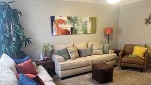 A living room with a beige sofa, a brown armchair, and a coffee table. There is a large abstract painting on the wall and a floor lamp. - NEW! Charming 2-bedroom townhome in convenient location (North Charleston)