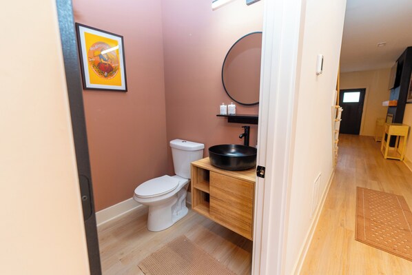 Un baño con inodoro, lavabo de color negro, espejo y repisa con artículos de aseo.