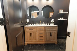 Un baño con un mueble de tocador de madera, dos espejos redondos y un estante con artículos de aseo.