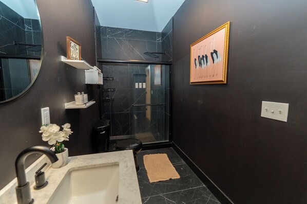 Un baño moderno con cabina de ducha de vidrio, paredes de mármol, un lavamanos blanco y una obra de arte enmarcada.