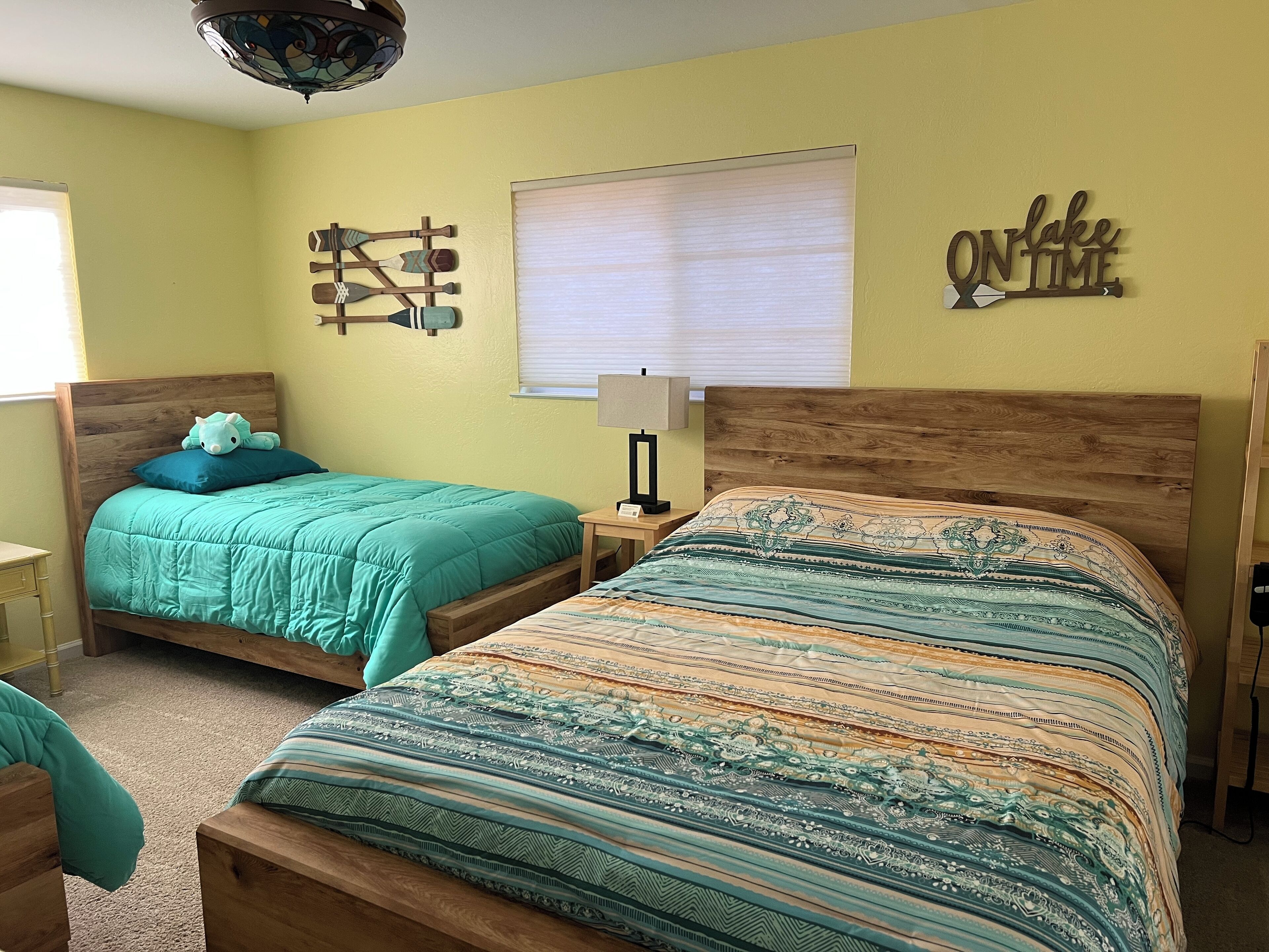 A bedroom with two beds, a window with blinds, a wall clock, and a decorative wall piece.