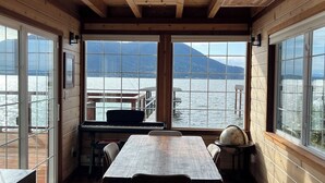 Ein Holzinnenraum mit einem großen Tisch und Stühlen, einem Klavier und großen Fenstern mit Blick auf einen See und Berge.