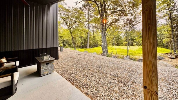A covered outdoor seating area with a stone fireplace, a wooden table, and a view of a forested landscape. - Couples Wilderness Suite (Cook)