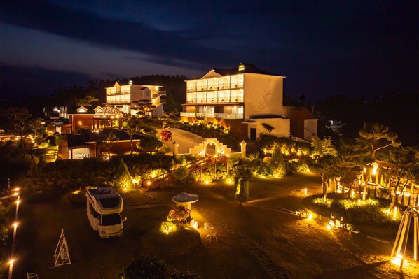 A multi-story building with a well-lit entrance and a parking area with a caravan. - Ocean The Hill (Taean)