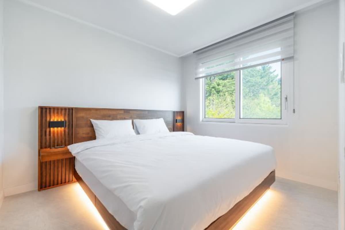 A modern bedroom with a wooden headboard, a large bed, and a window with a view of greenery.