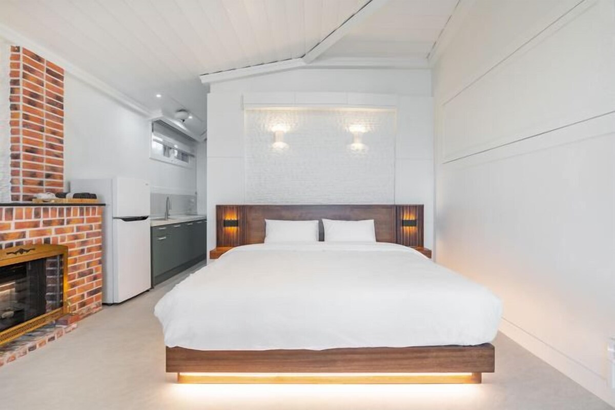 A modern bedroom with a large bed, a brick fireplace, and a kitchen area in the background.