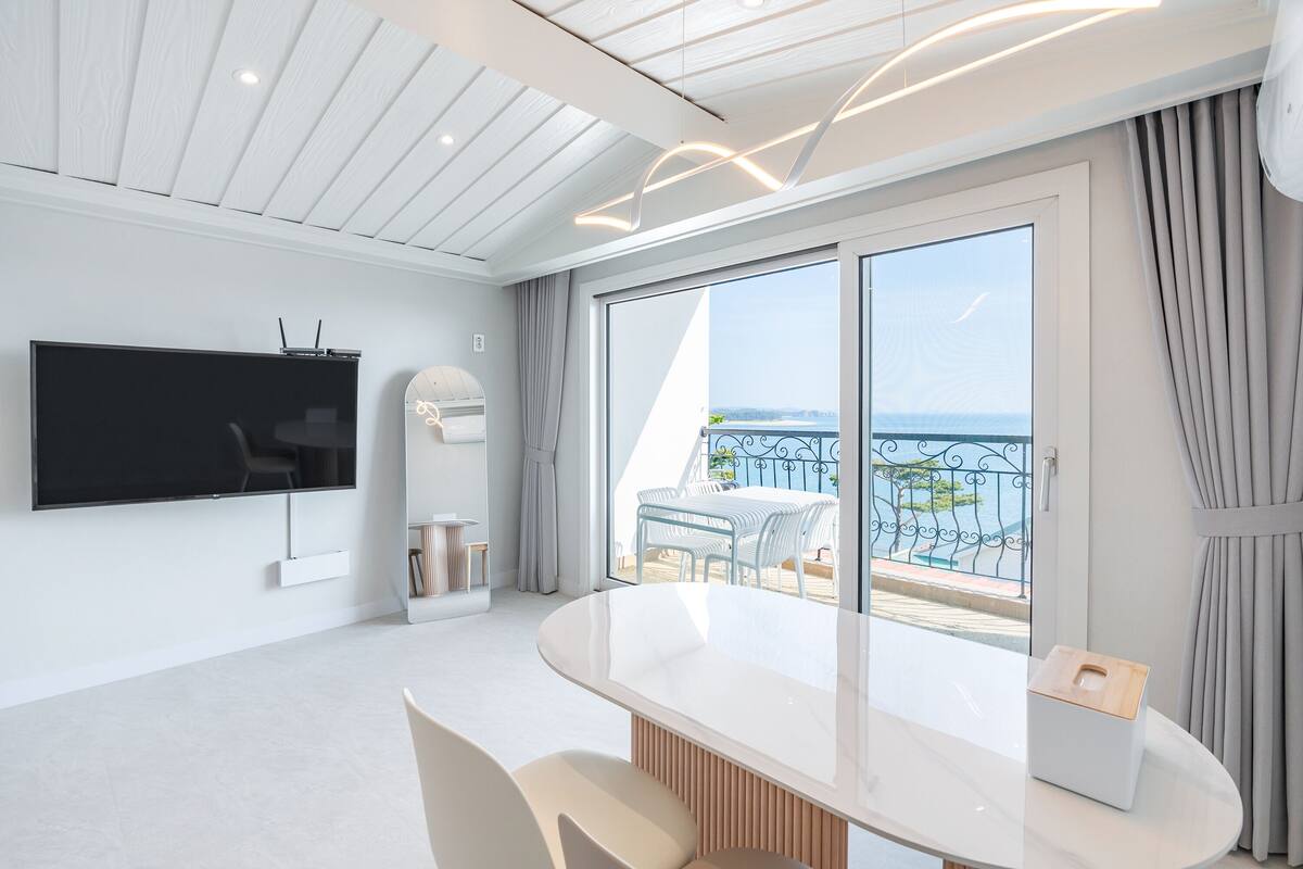 A modern room with a large TV, a round table, and a balcony with a view of the sea.