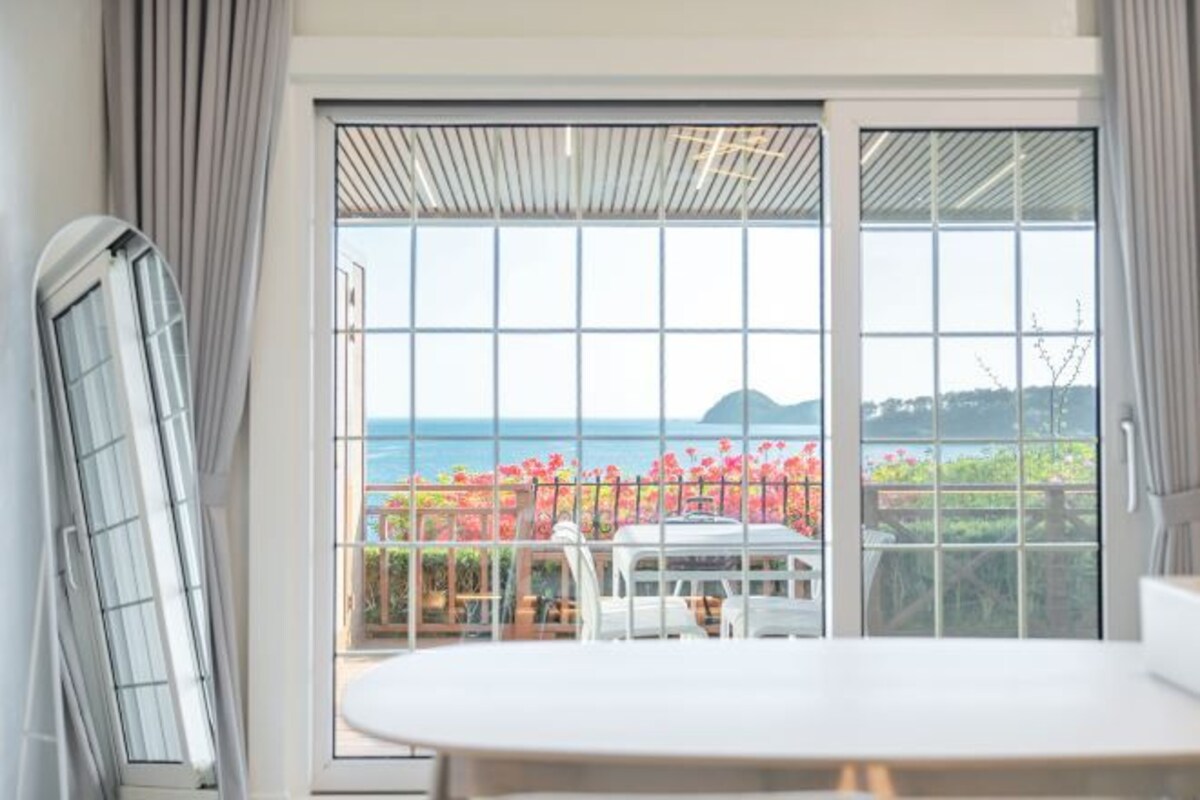 A room with a large sliding glass door leading to a balcony with a view of the sea and mountains.