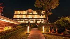 A multi-story hotel with a balcony on each floor, a wooden pathway leading to the entrance, and a large tree in the foreground. - Ocean The Hill (Taean)
