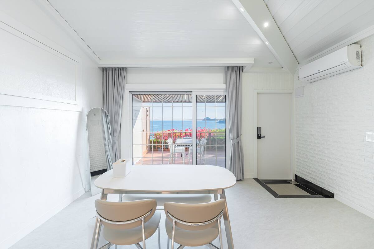 A modern room with a white table and chairs, a large window with a view of the sea, and a white brick wall.