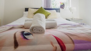 A neatly made bed with a pillow, a rolled towel, and a blanket with a face design. - WoodHus Lodge  Large Double Room 3 (Stoke-on-Trent)