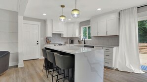 A modern kitchen with white cabinetry, a large island with bar stools, pendant lighting, and a view of the outdoors. - Zest of Zionsville with WiFi, AC (Zionsville)