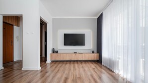 Una sala de estar moderna con un televisor de pantalla plana grande montado en la pared, un mueble de madera debajo y un ventanal con cortinas translúcidas.
