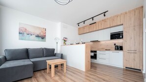 Una sala de estar moderna con un sofá gris, una pequeña mesa de madera y una zona de cocina con armarios blancos y detalles de madera.