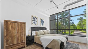 Une chambre moderne avec une grande fenêtre, une armoire en bois, un lit recouvert d’une couette blanche et deux tableaux encadrés accrochés au mur.