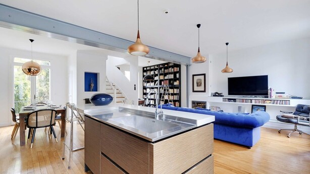 Una cocina moderna con una isla central, una zona de comedor con mesa y sillas, un sofá azul y una estantería al fondo.