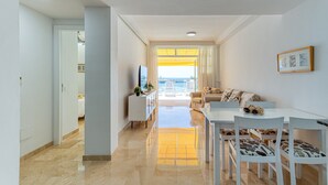 A modern living room with a sofa, dining table, and chairs. There is a TV on a cabinet, and a window with curtains. - Apartment in Playa de Mogán by the Beach (Mogán)