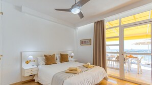 A hotel room with a bed, nightstands, a ceiling fan, a window with curtains, and a view of the outside. - Apartment in Playa de Mogán by the Beach (Mogán)