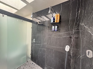 Moderni kylpyhuone, jossa on tummat marmoriseinät, lasinen suihkuseinä ja kaksi hyllyä, joilla on hygieniatuotteita.