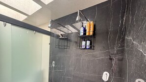 Une salle de bain moderne avec des murs en marbre foncé, une douche en verre et deux étagères où sont rangés les articles de toilette.
