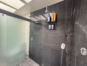 A modern bathroom with dark marble walls, a glass shower enclosure, and two shelves holding toiletries. - Departamento Miles Davis (Val'Quirico)