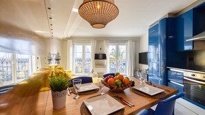 A modern kitchen with blue cabinets, a large island with a fruit bowl, and a dining area with a view.