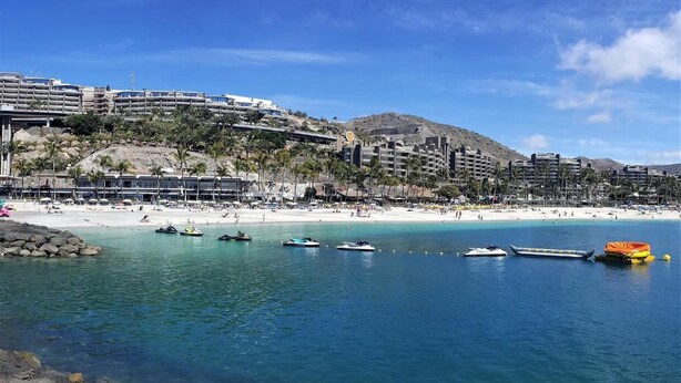 A coastal resort with a beach, clear blue waters, and a mountainous backdrop.