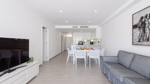 A modern living room with a flat-screen TV, a grey sofa, a dining table with chairs, and a kitchen in the background. - Apartment in Playa de Mogán with Sea Views (Mogán)